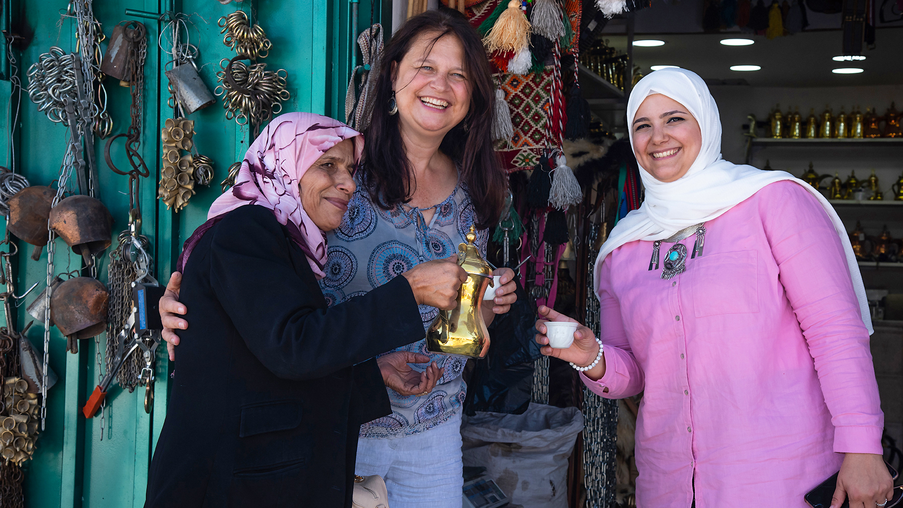 Drei Frauen, eine mit Teekanne und Tasse, lachen vor einem Marktstand voller Metallwaren.