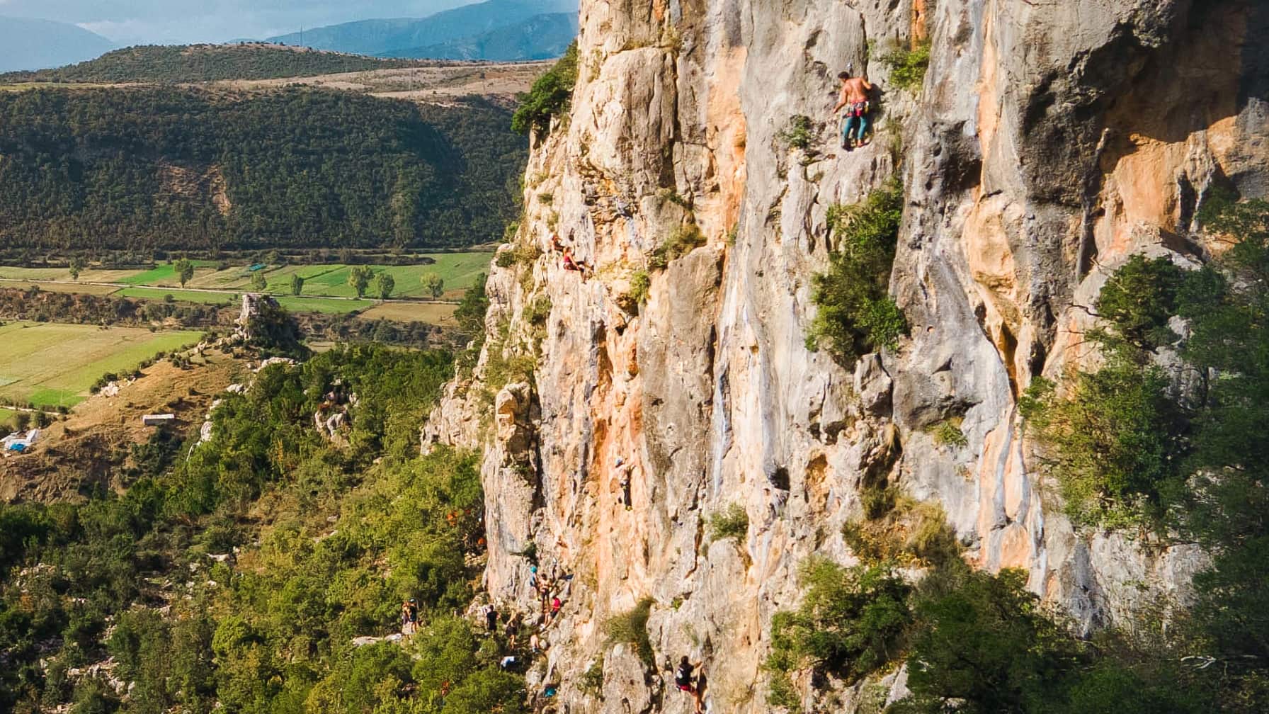 Kletterer erklimmen eine steile, felsige Klippe mit einer weiten grünen Landschaft im Hintergrund.