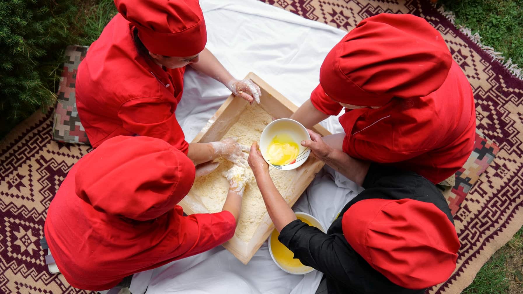 Vier Frauen in roten Kopfbedeckungen kneten Teig in einer Holzkiste auf einem traditionellen Teppich