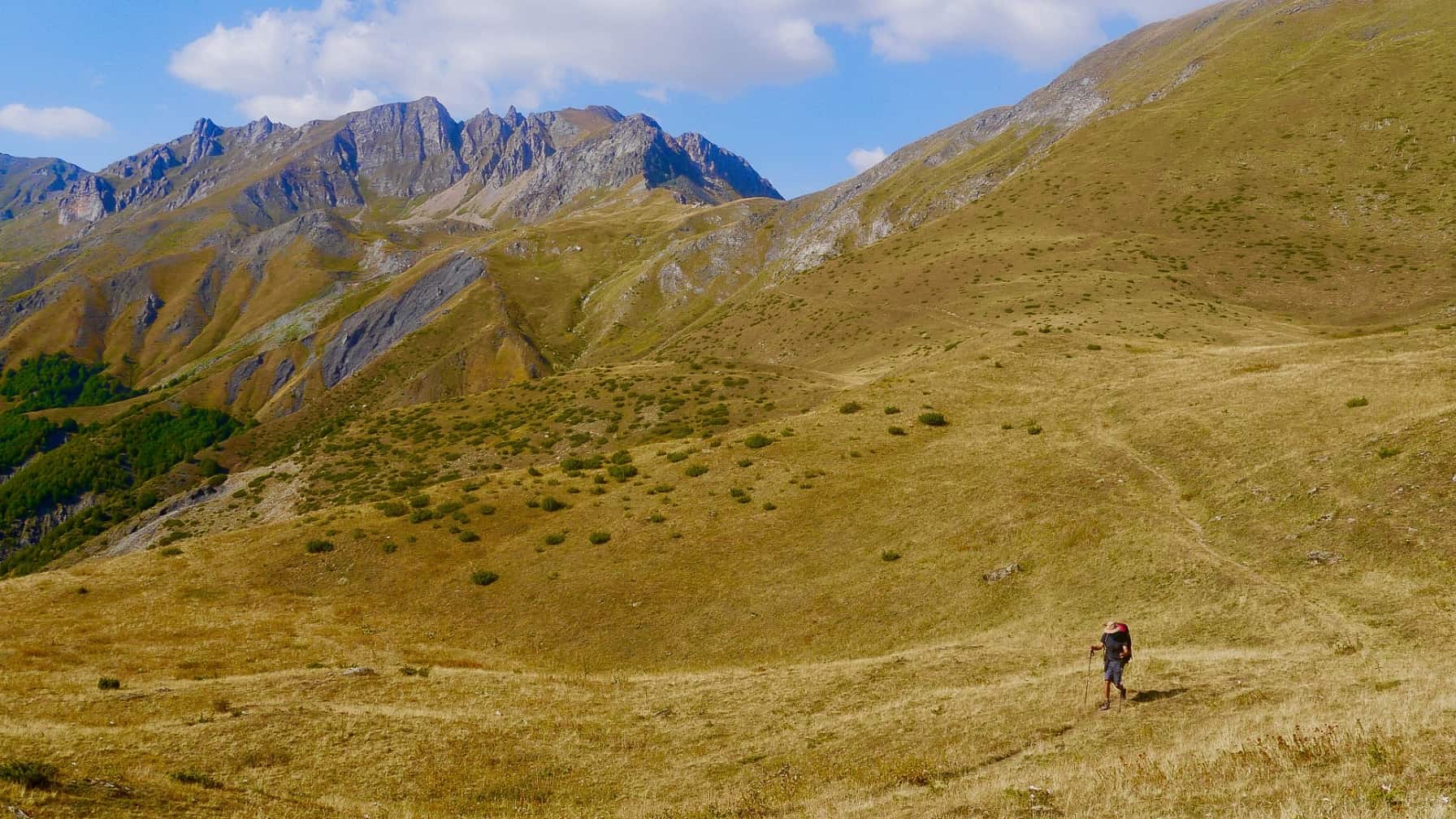 Ein Wanderer mit Rucksack steigt eine goldene Hügellandschaft hinauf, mit zerklüfteten Bergen im Hintergrund.