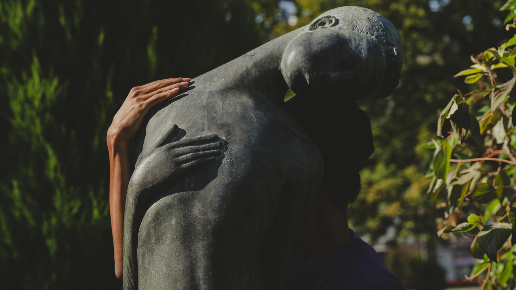 Eine echte Person umarmt eine weibliche Bronzestatue, wobei die menschliche Hand und die Statuehand parallel auf dem Rücken liegen.
