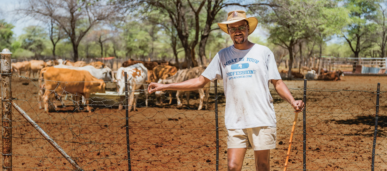 Namibischer Bauer vor einem Zaun stehend. Er trägt ein T-Shirt, eine helle Hose und einen Hut und schaut in die Kamera. Im Hintergrund sind unscharf Kühe und eine ländliche Umgebung zu erkennen.