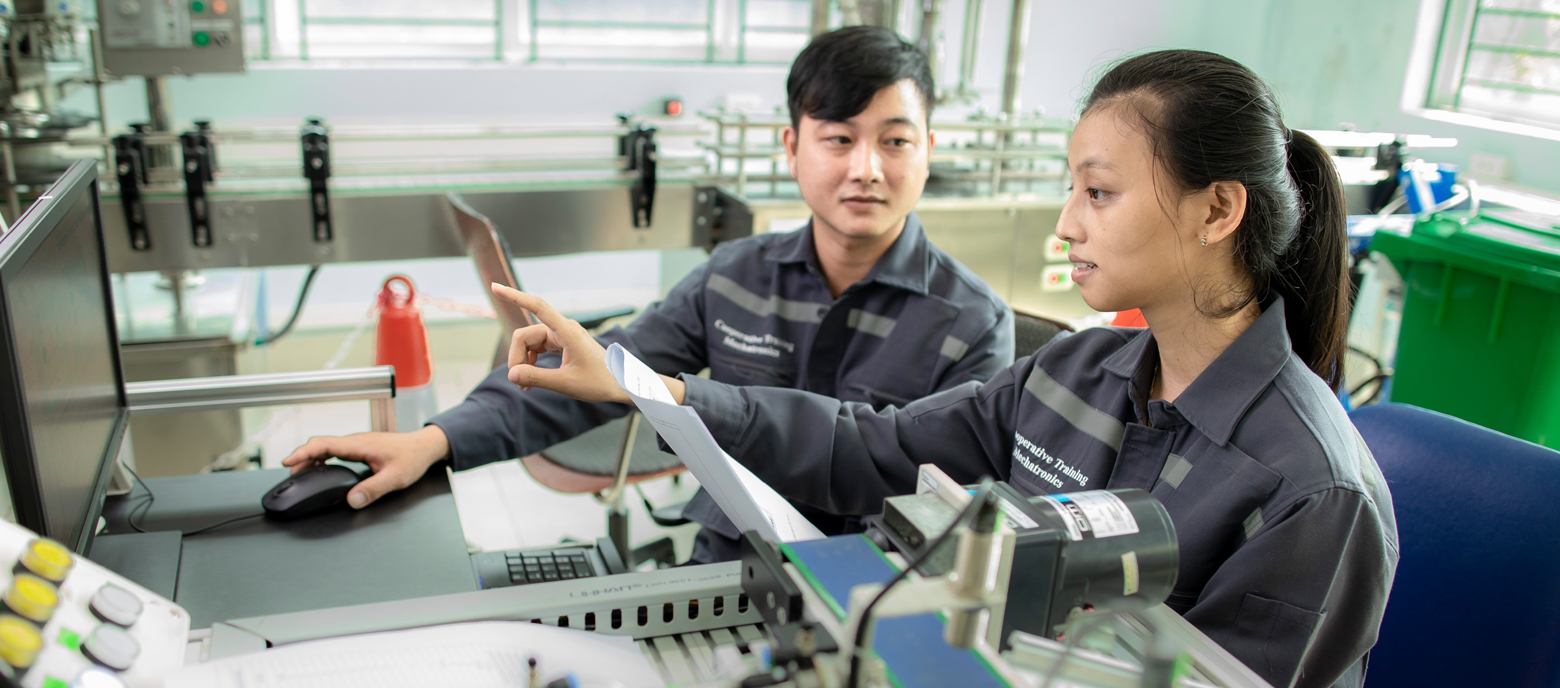 Das Foto zeigt einen Mann und eine Frau in Arbeitskleidung in Vietnam, die gemeinsam vor einem Computer und weiterer Technik sizen. Sie tragen beide graue Overalls und stehen vor einem Arbeitstisch mit verschiedenen technischen Geräten.