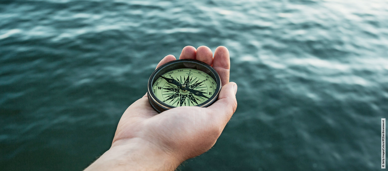 Nahaufnahme einer Hand, die einen Kompass vor verschwommenem Meer im Hintergrund hält.