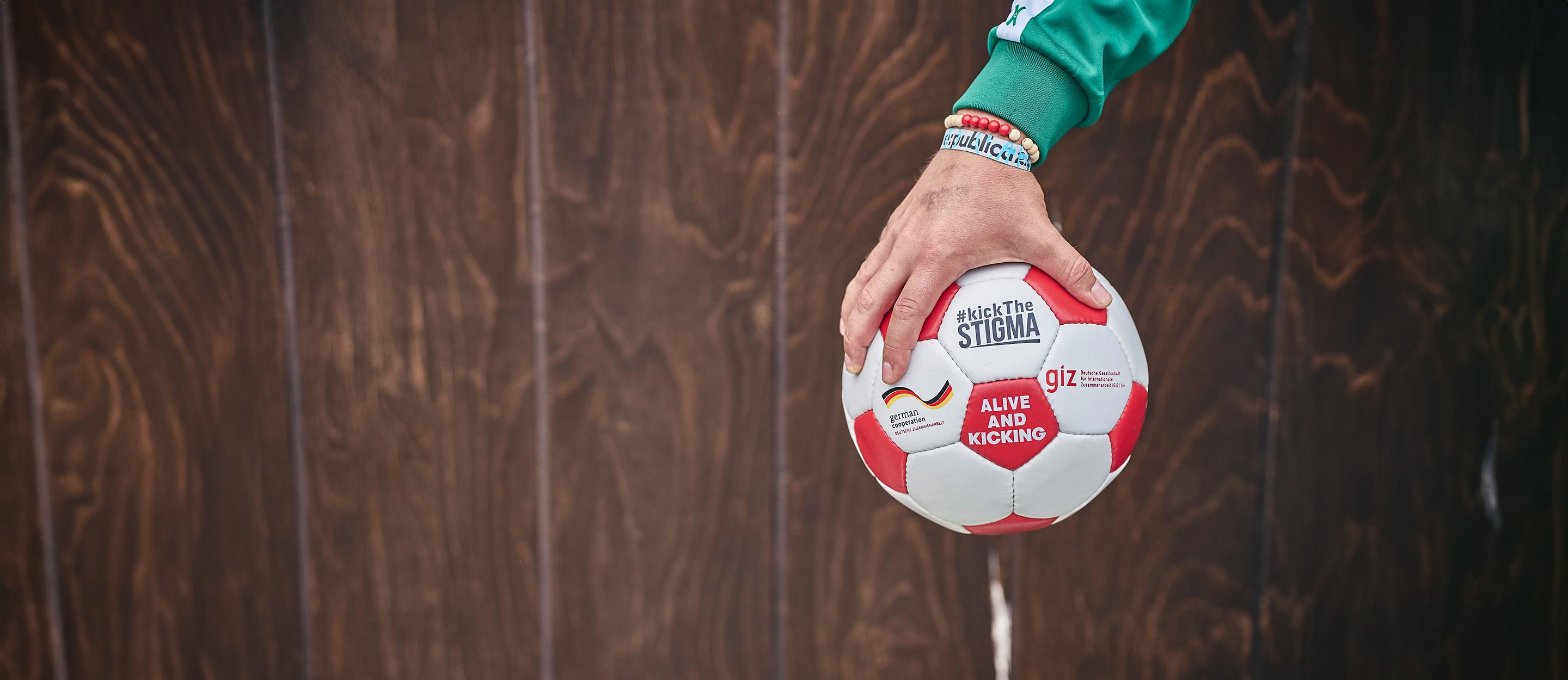 Eine Hand, die einen Fußball ins Bild hält. Aufschrift: #kickTheSTIGMA