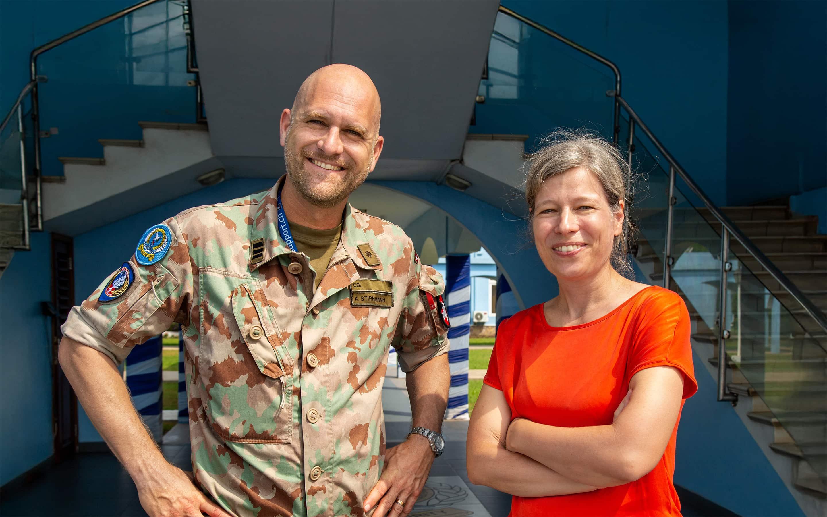 Ein lachender Mann in einer militärischen Tarnuniform mit Namensschild „A. Stirnimann“ steht neben einer Frau in einem roten Shirt vor einer modernen blauen Gebäudestruktur.