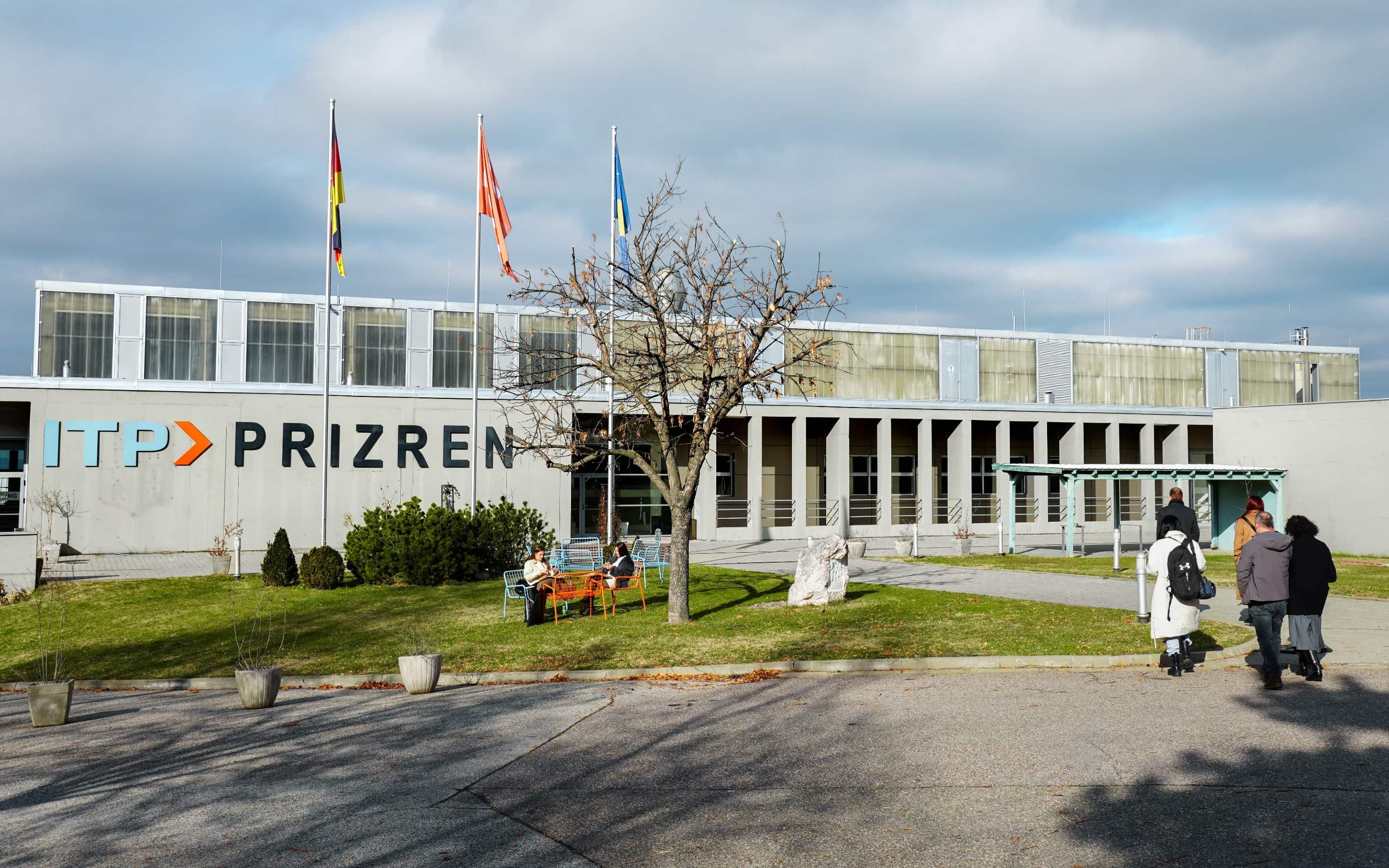 Außenansicht des ITP Prizren-Gebäudes mit drei Fahnenmasten, Sitzgelegenheiten auf der Wiese und mehreren Personen auf dem Gelände.