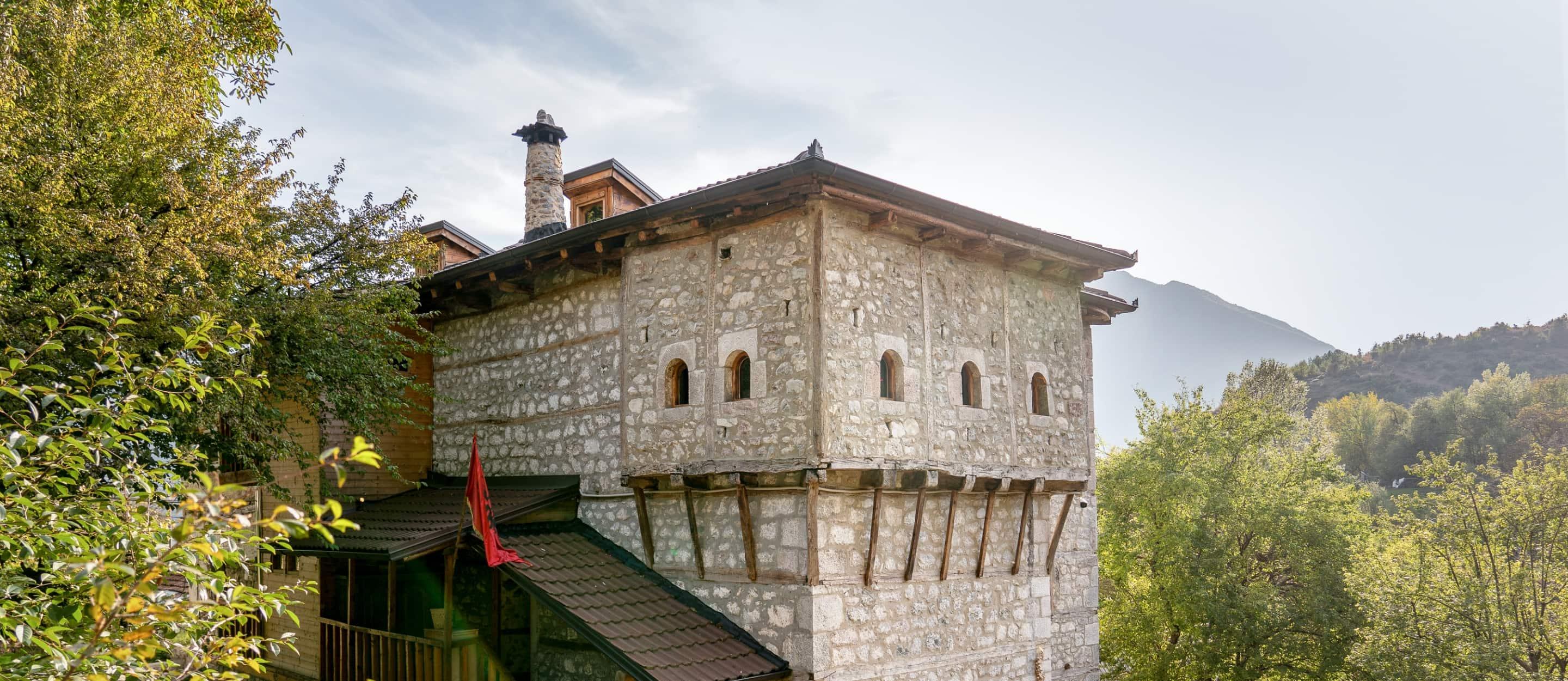 Ein traditionelles Steinhaus mit kleinen Fenstern, Holzdetails und der roten Fahne Albaniens steht inmitten grüner Bäume mit Bergen im Hintergrund.