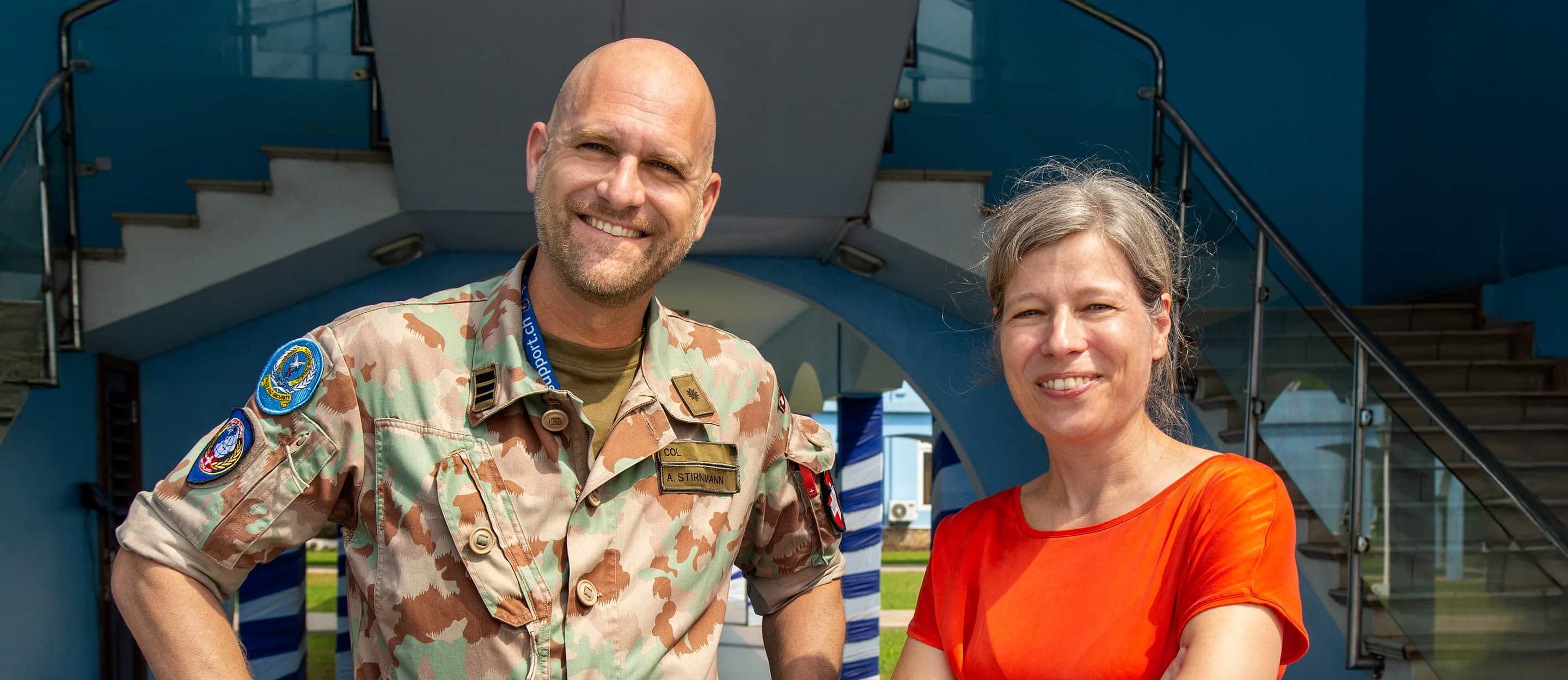 Ein lachender Mann in einer militärischen Tarnuniform mit Namensschild „A. Stirnimann“ steht neben einer Frau in einem roten Shirt vor einer modernen blauen Gebäudestruktur.