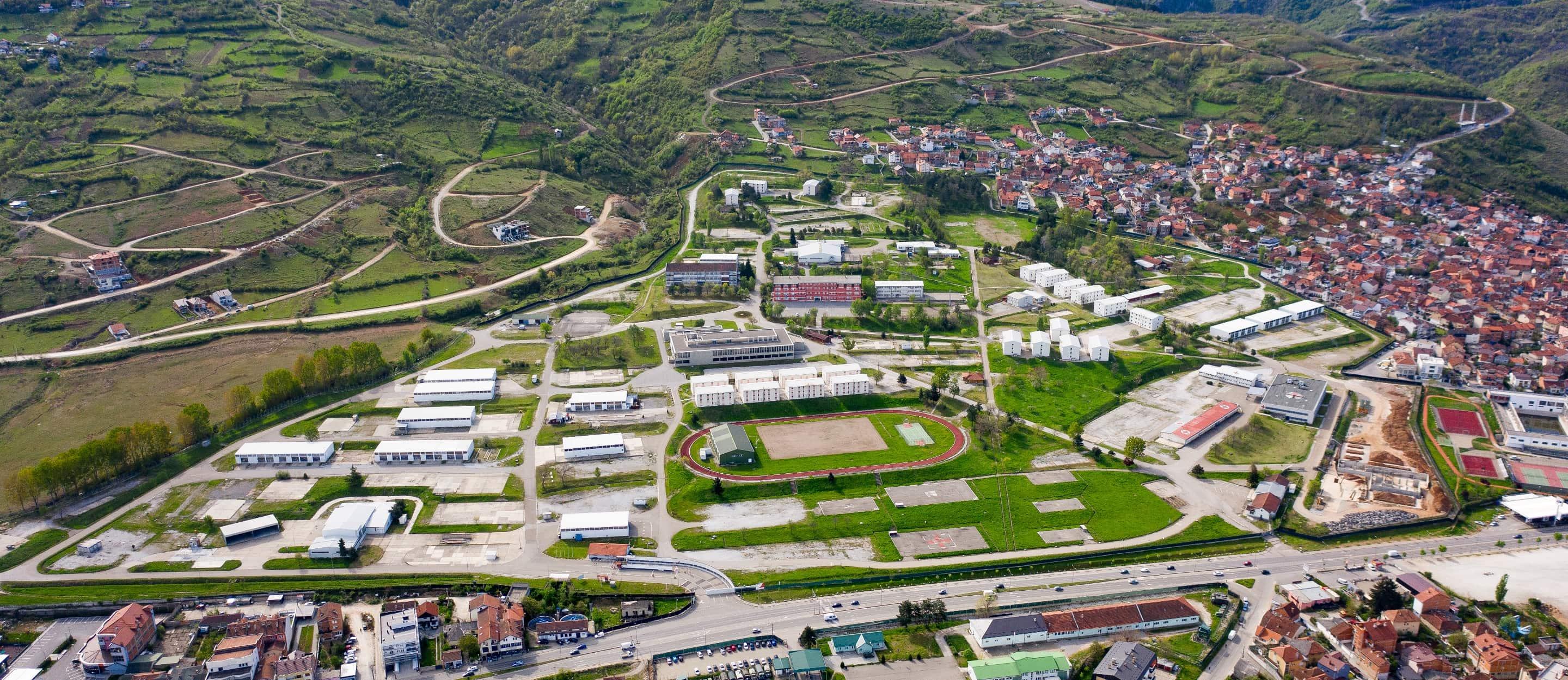 Luftaufnahme eines weitläufigen Geländes mit Gebäuden, Sportanlagen und einer angrenzenden Stadt in einer hügeligen Landschaft.