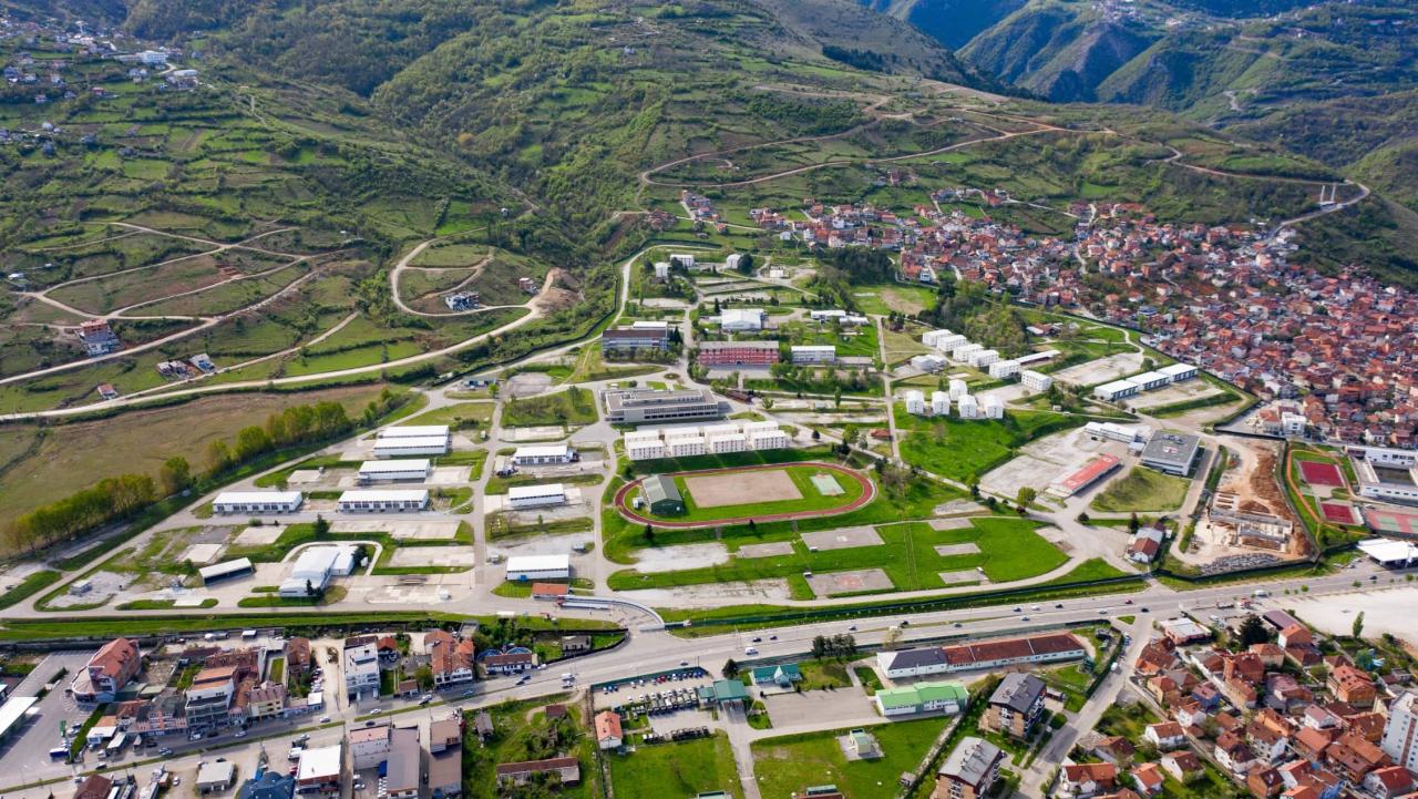 Luftaufnahme eines weitläufigen Geländes mit Gebäuden, Sportanlagen und einer angrenzenden Stadt in einer hügeligen Landschaft.