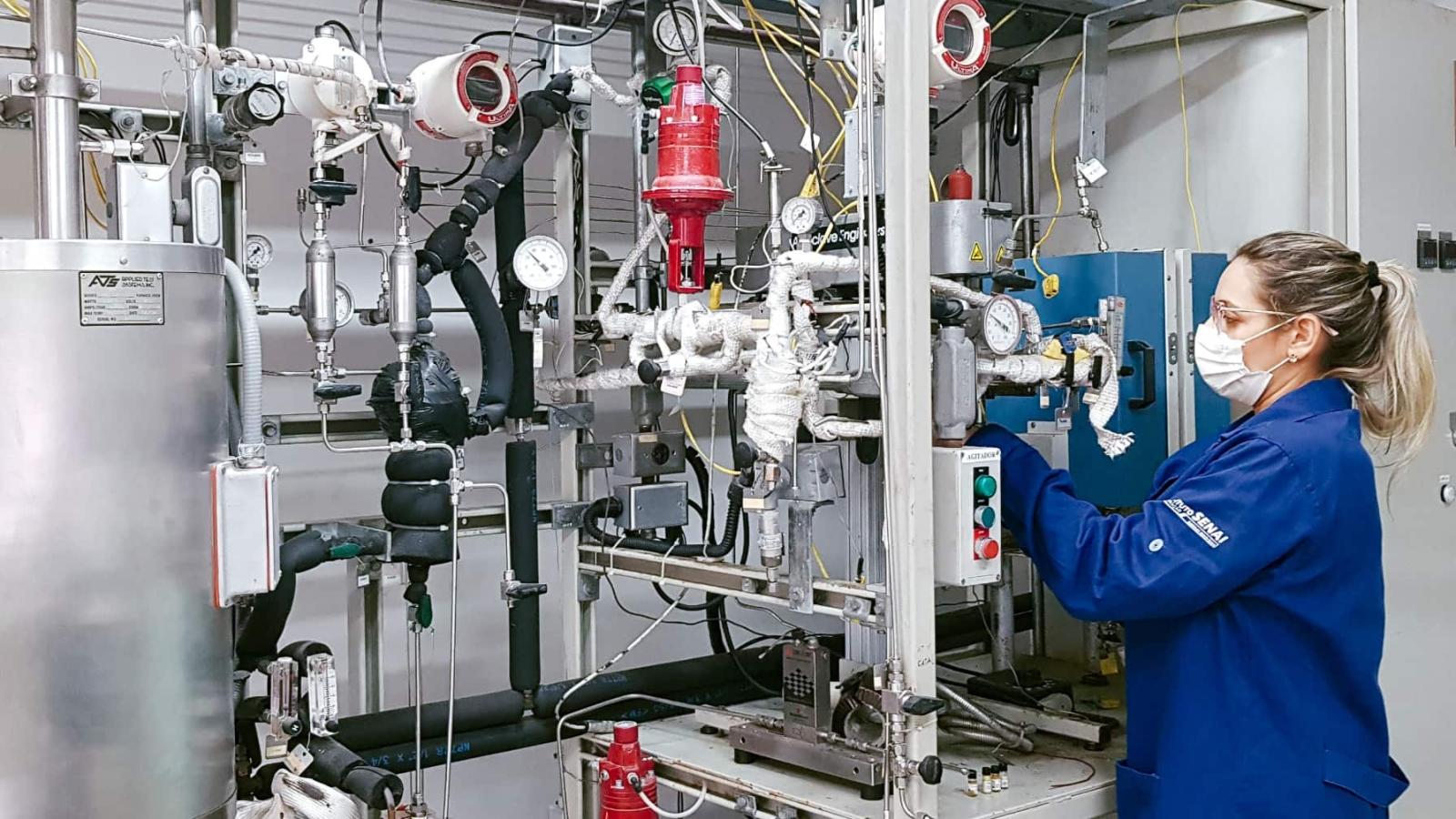 Renata Moura/GIZ Brasil Eine Frau in einem blauen Laborkittel und einer Gesichtsmaske arbeitet an einer komplexen, industriellen Apparatur mit zahlreichen Rohren, Anzeigen und Ventilen.