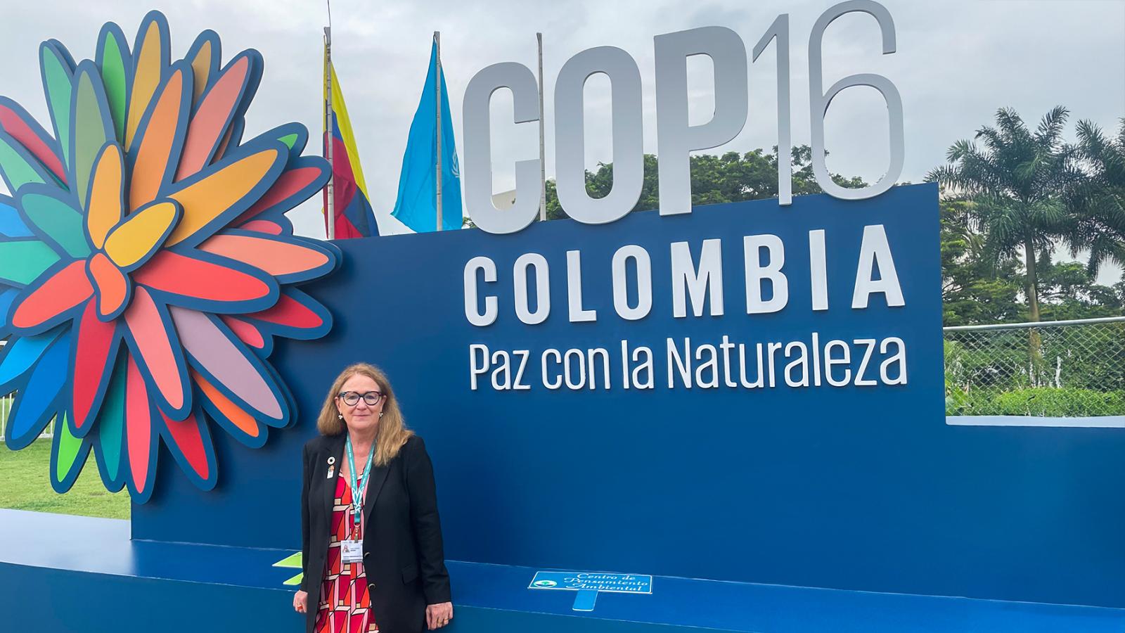 Eine Frau steht vor einem Schild mit der Aufschrift "COP16 Colombia Paz con la Naturaleza" im Freien, trägt eine schwarze Jacke und ein gemustertes Kleid.