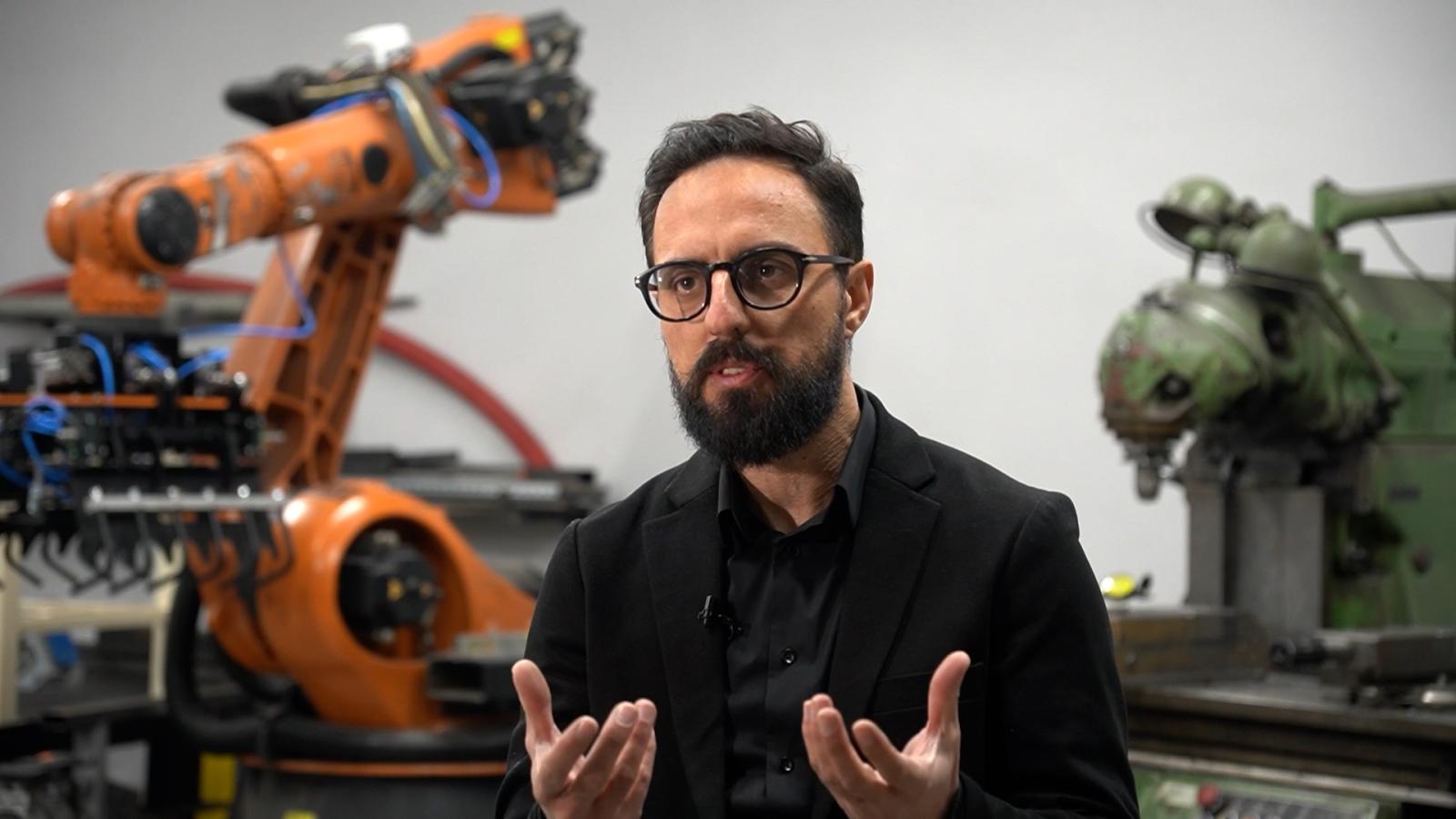 Ein Mann mit Brille und dunklem Bart spricht gestikulierend in einem industriellen Umfeld mit einem orangefarbenen Roboterarm und einer grünen Maschine im Hintergrund.