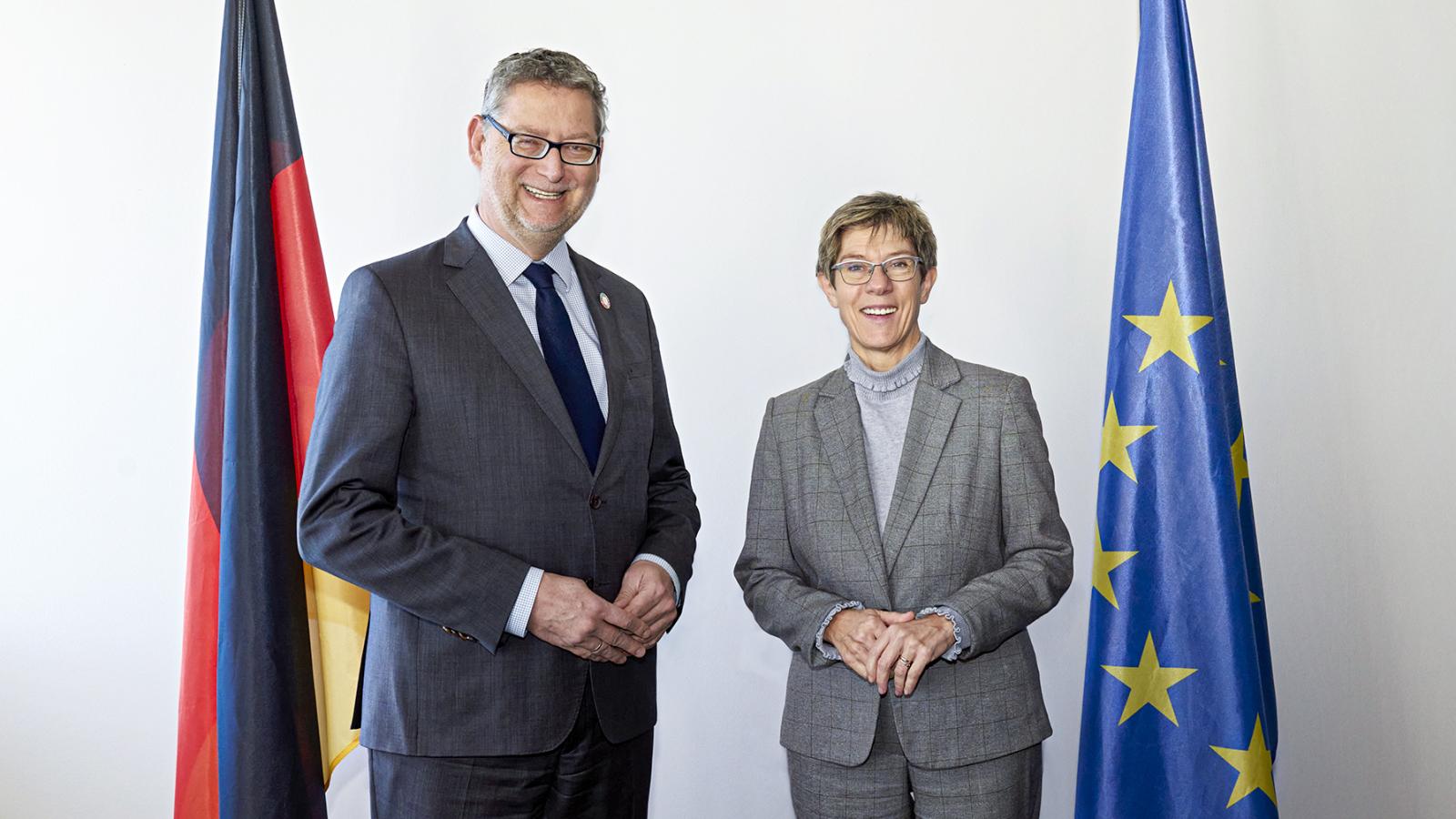 Gaby Gerster/GIZ Zwei Personen in formeller Kleidung stehen nebeneinander und lächeln in die Kamera, flankiert von einer Deutschlandflagge links und einer Europaflagge rechts auf hellem Hintergrund.