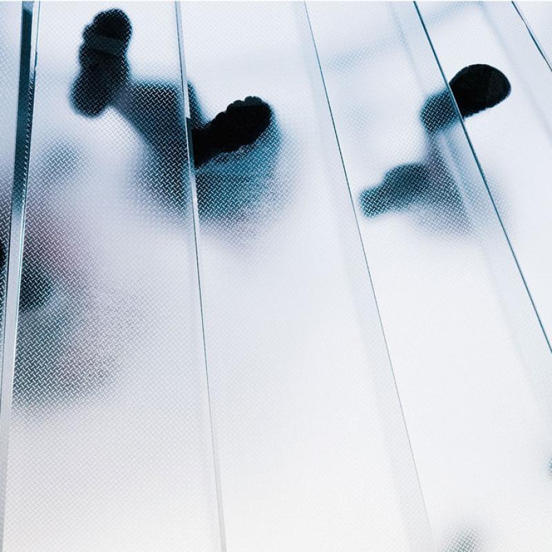 Das Foto zeigt die Schatten mehrerer Personen, die über eine durchscheinende Glas- oder Kunststofffläche laufe. Die Oberfläche ist in Streifen unterteilt, was die Schatten zusätzlich streckt und ein dynamisches Muster erzeugt. Die Perspektive ist von unten nach oben gerichtet, wobei die Füße und Beine der Personen deutlich erkennbar sind.