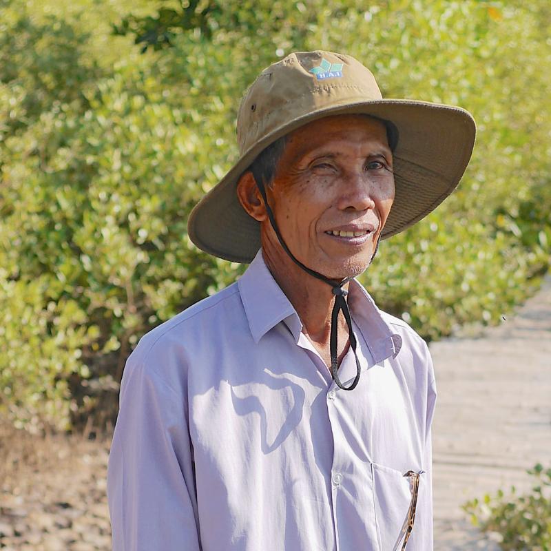 Das Bild zeigt einen älteren Mann, der in einer natürlichen Umgebung steht. Er trägt einen hellen Hut, ein weißes Hemd und blickt in die Ferne. Der Hintergrund zeigt einen trockenen Boden und dichtes Buschwerk entlang eines schmalen Weges.