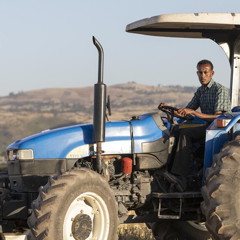 Ein Bauer in Äthiopien fährt einen blauen Traktor über ein trockenes Feld mit Hügeln und einer kleinen Siedlung im Hintergrund. Der Himmel ist klar, und der Bauer schaut konzentriert in die Kamera.