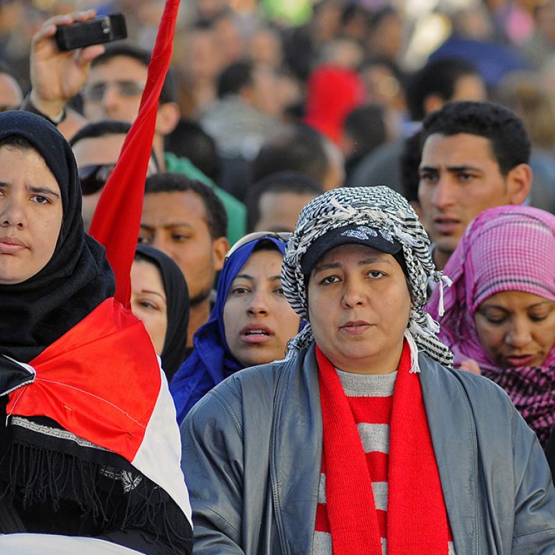 Zwei Frauen, eine mit einem schwarzen Kopftuch und die andere mit einer grauen Strickmütze und einem roten Schal, tragen eine Fahne. Eine dritte Person hebt ein rotes Tuch hoch. Im Hintergrund ist eine Menschenmenge zu sehen.