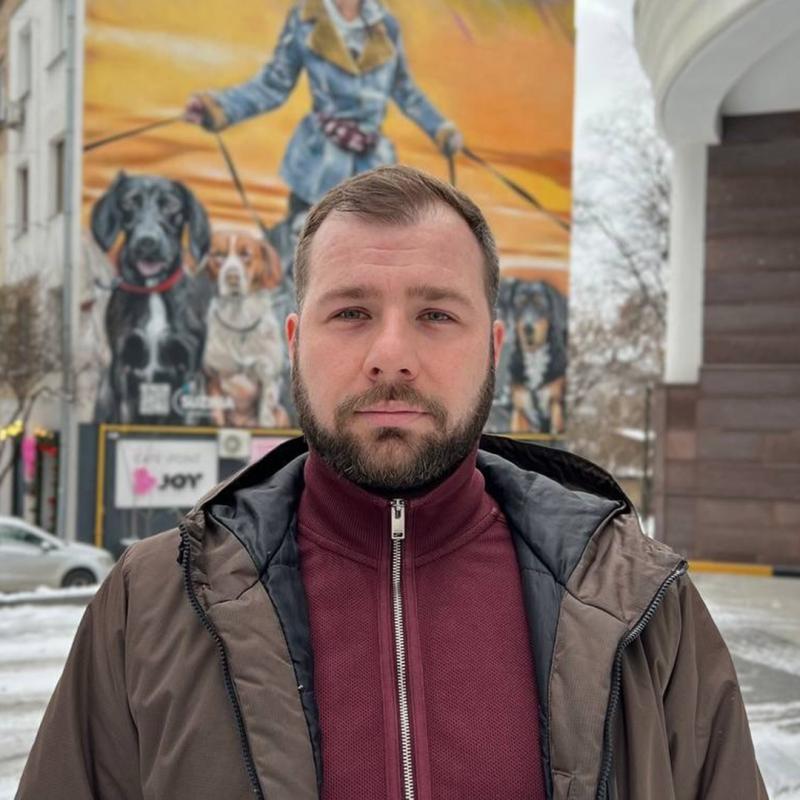 Ein Mann mit kurzem Bart und brauner Jacke steht vor einer bunten Wandmalerei, die eine Person mit mehreren Hunden an der Leine zeigt.