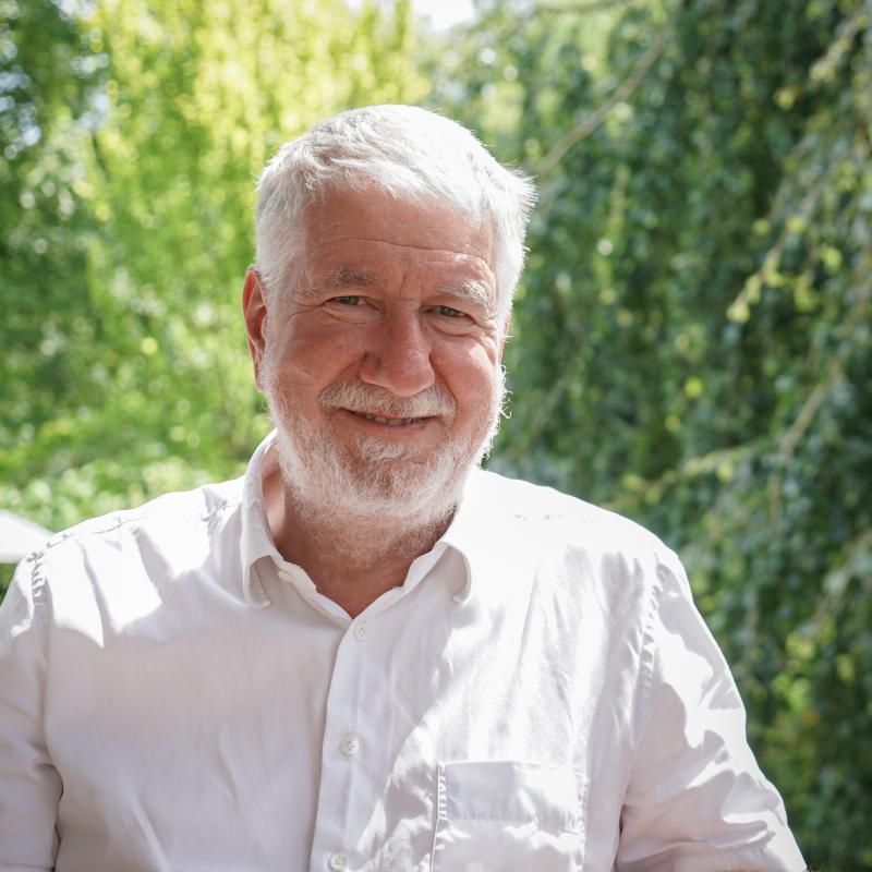 Ein älterer Mann mit weißem Haar und Bart lächelt in einem weißen Hemd vor einer sonnigen, grünen Naturkulisse in die Kamera.