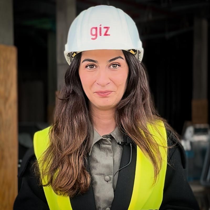 Eine Frau mit langen braunen Haaren trägt einen weißen Schutzhelm mit der Aufschrift "giz" und eine gelbe Warnweste auf einer Baustelle.