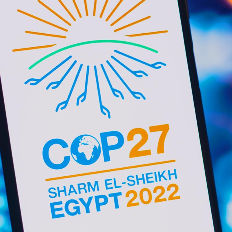 Ein Werbeplakat für die Klimakonferenz COP27 in Sharm El-Sheikh, Ägypten 2022. Das Design zeigt eine stilisierte Sonne in Orange und Grün über dem blauen Logo für COP27, darunter der Globus, und den Veranstaltungsort und das Jahr in Blau und Orange.