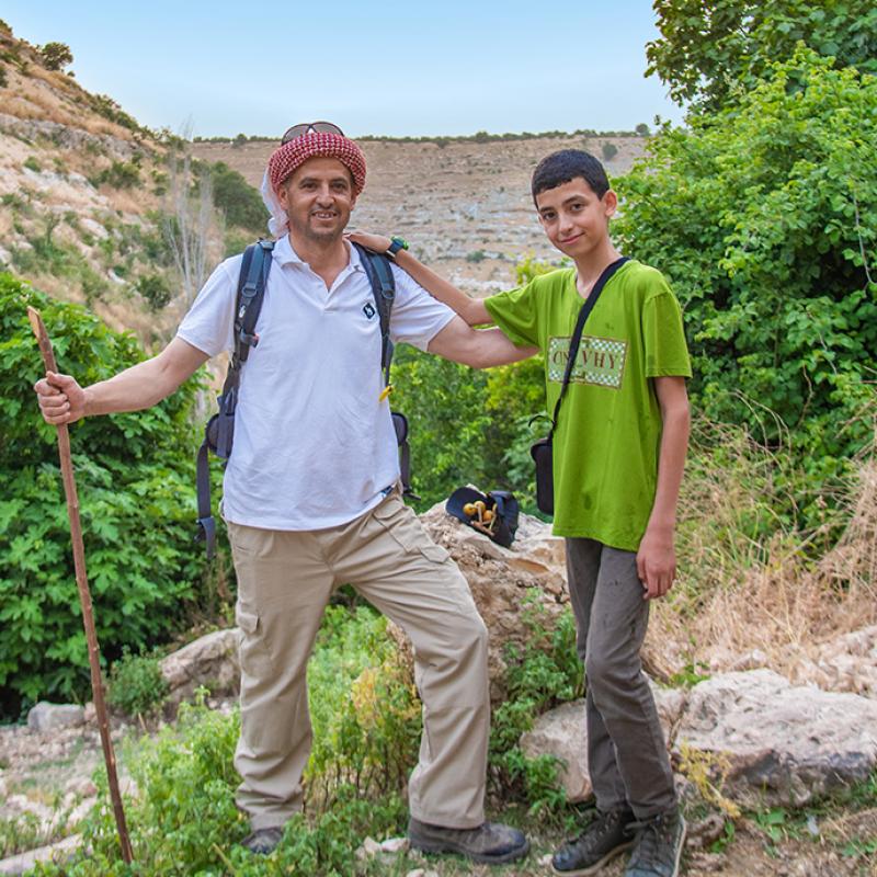 Ein erwachsener Mann und ein Jugendlicher stehen auf einem Naturpfad in Jordanien. Der Mann trägt eine rote Kopfbedeckung, ein weißes Hemd, eine beige Hose und hält einen Wanderstab; der Junge trägt ein grünes Shirt und graue Hose. Sie lächeln beide und stehen vor einer hügeligen, bewachsenen Landschaft.