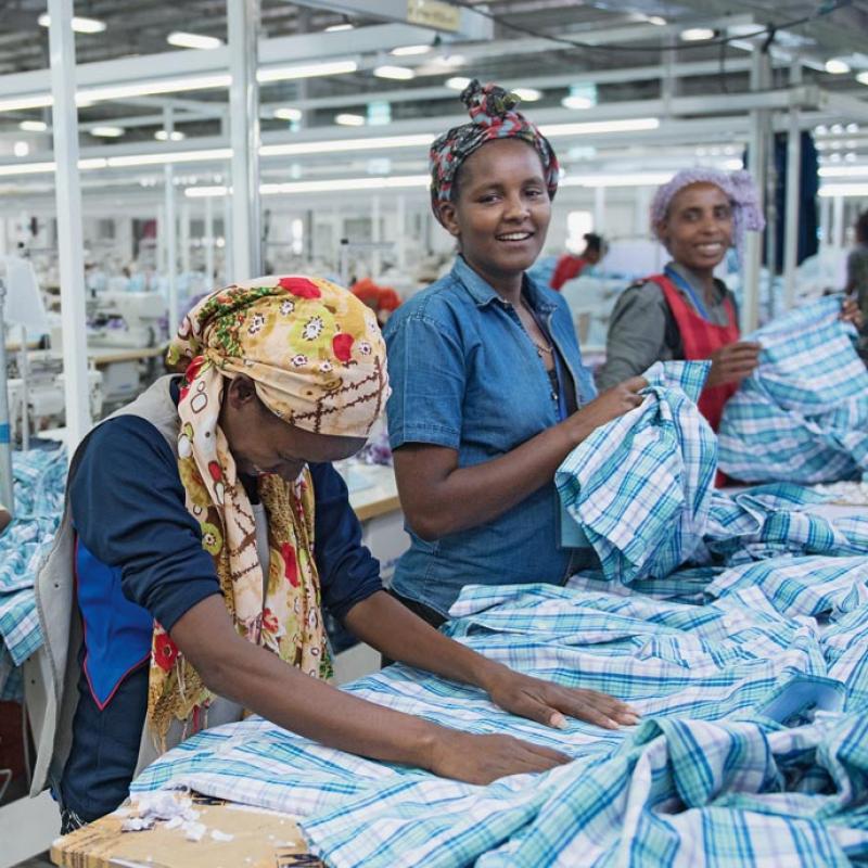 Drei Frauen arbeiten in einer Textilfabrik in Äthopien. Sie stehen an Tischen mit Stapeln von blau-weiß karierten Stoffen. Die Frau im Vordergrund trägt ein gelbes Kopftuch, die mittlere Frau ein blaues Oberteil und die dritte Frau im Hintergrund ein rotes. Im Hintergrund sind weitere Arbeitsplätze und Personen sichtbar.