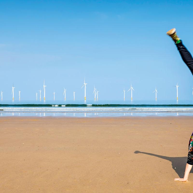 Foto einer Person, die einen Handstand mit gespreizten Beinen auf einem Strand ausführt, im Hintergrund sind mehrere Windturbinen auf dem Meer. Die Person ist von der Kamera weggerichtet, sodass das Gesicht nicht sichtbar ist, und trägt bunte Kleidung.