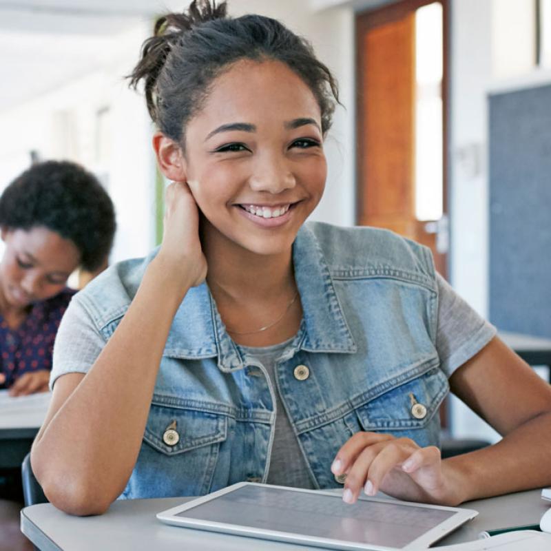 Junge Schülerin mit einem Tablet lächelt in die Kamera, während sie am Tisch in einem Klassenzimmer sitzt. Im verschwommenem Hintergrund arbeiten andere Schüler konzentriert und ein Lehrer geht durch den Raum.