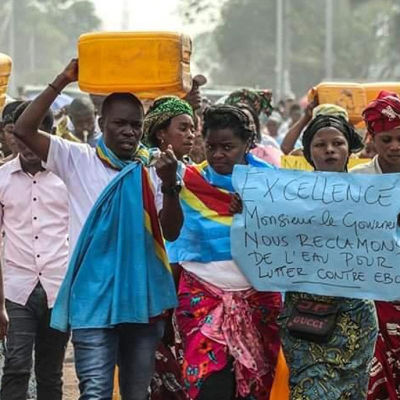 Eine Gruppe von Menschen marschiert auf einer staubigen Straße im Kongo, viele tragen gelbe Wasserkanister auf dem Kopf. Eine Person hält ein Plakat hoch, auf dem auf Französisch steht: Exzellenz, Herr Gouverneur, wir fordern sauberes Wasser, um gegen Ebola zu kämpfen. Einige tragen traditionelle, bunte Kleidung, andere einfache Alltagskleidung.