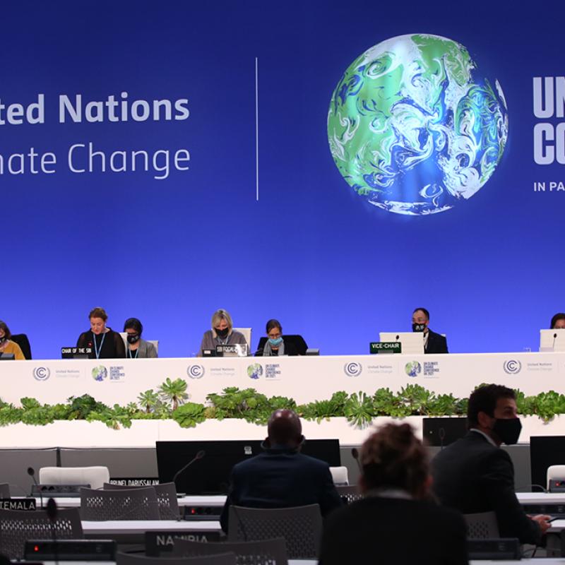 Blick auf das Podium der UN-Klimakonferenz 2021 im Vereinigten Königreich. Delegierte sitzen hinter einem langen Tisch mit Namensschildern und grüner Dekoration. Im Hintergrund sind das Logo der Vereinten Nationen zu sehen und im Vordergrund das Publikum, das den Rednern zuhört.