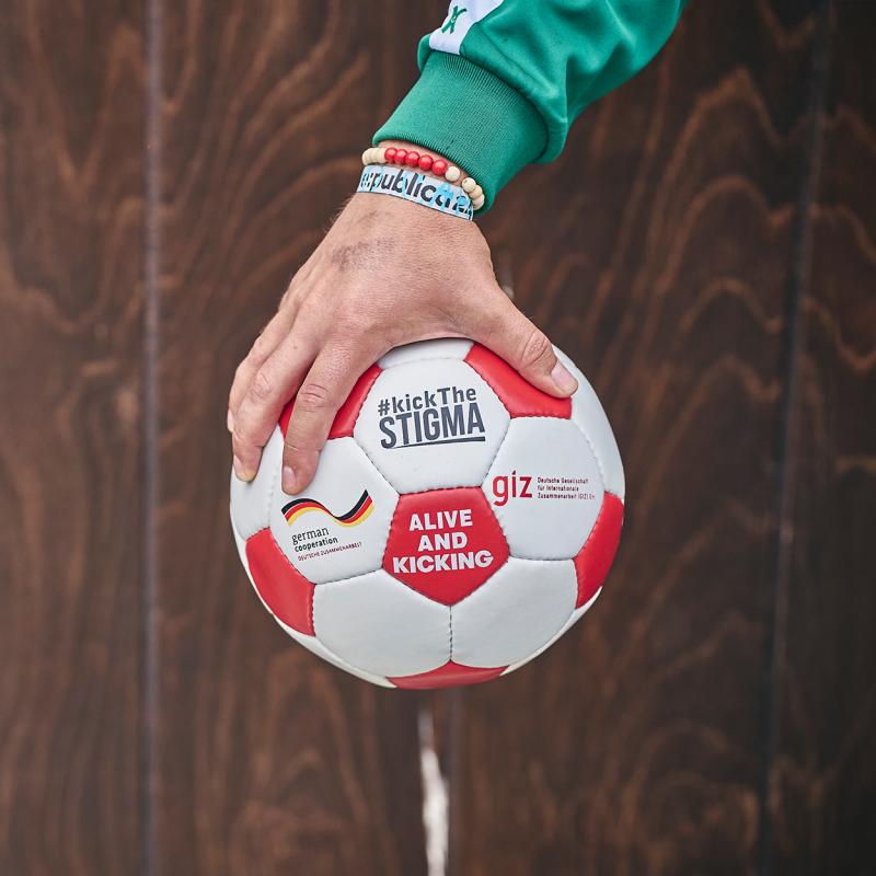 Eine Hand, die einen Fußball ins Bild hält. Aufschrift: #kickTheSTIGMA