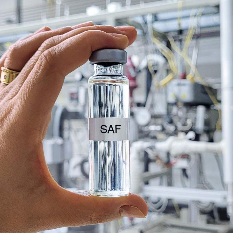 Eine Hand hält ein kleines, mit "SAF" beschriftetes Fläschchen mit klarer Flüssigkeit vor einem verschwommenen industriellen Hintergrund.