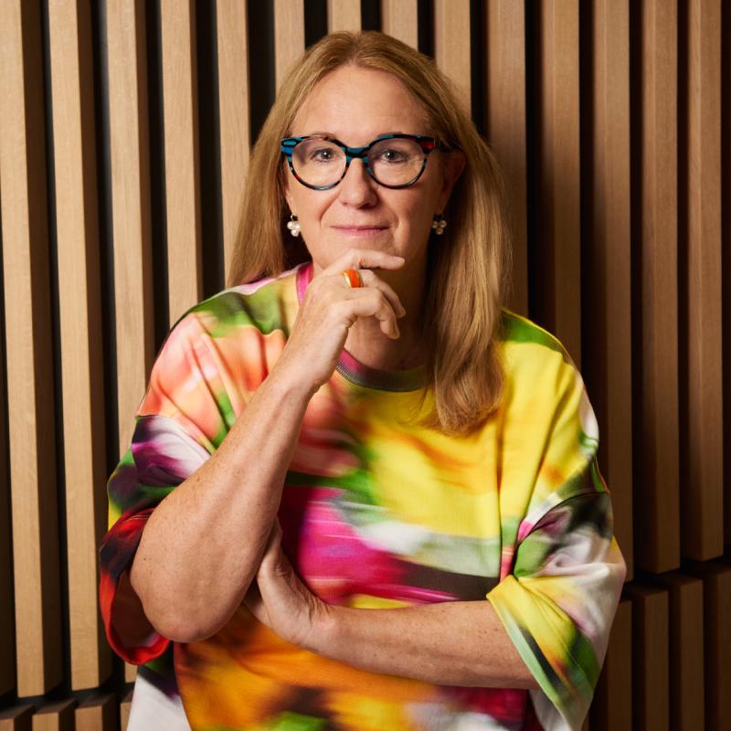 Eine Frau mit langen blonden Haaren und Brille steht vor einer Wand mit Holzlamellen und trägt eine bunte Bluse.