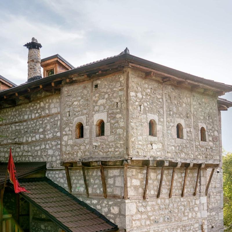 Ein traditionelles Steinhaus mit kleinen Fenstern, Holzdetails und der roten Fahne Albaniens steht inmitten grüner Bäume mit Bergen im Hintergrund.
