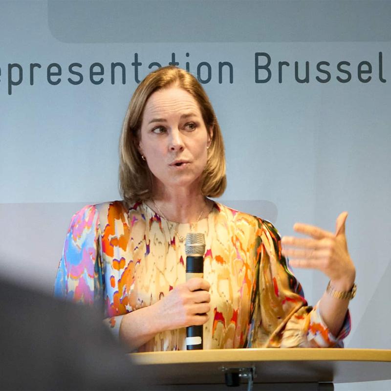  Eine Frau mit schulterlangem blondem Haar trägt eine farbenfrohe Bluse mit abstraktem Muster und spricht auf einer Veranstaltung in Brüssel an einem Rednerpult mit einem Mikrofon in der Hand.