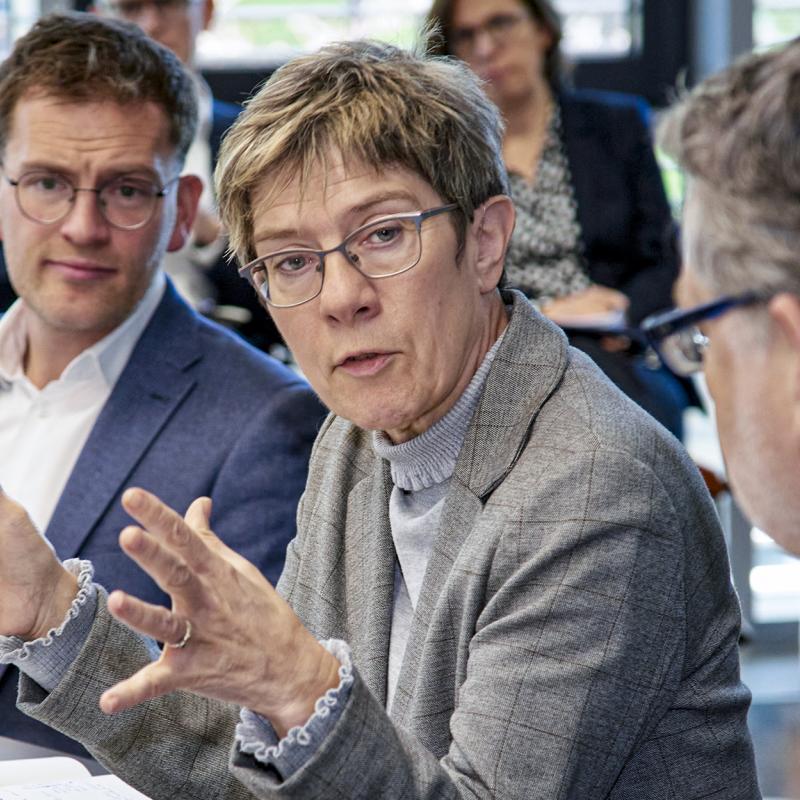 Diskussion in einem Konferenzraum mit einer Frau im grauen Blazer, die mit Gestik spricht, umgeben von mehreren Personen in Businesskleidung, die aufmerksam zuhören.