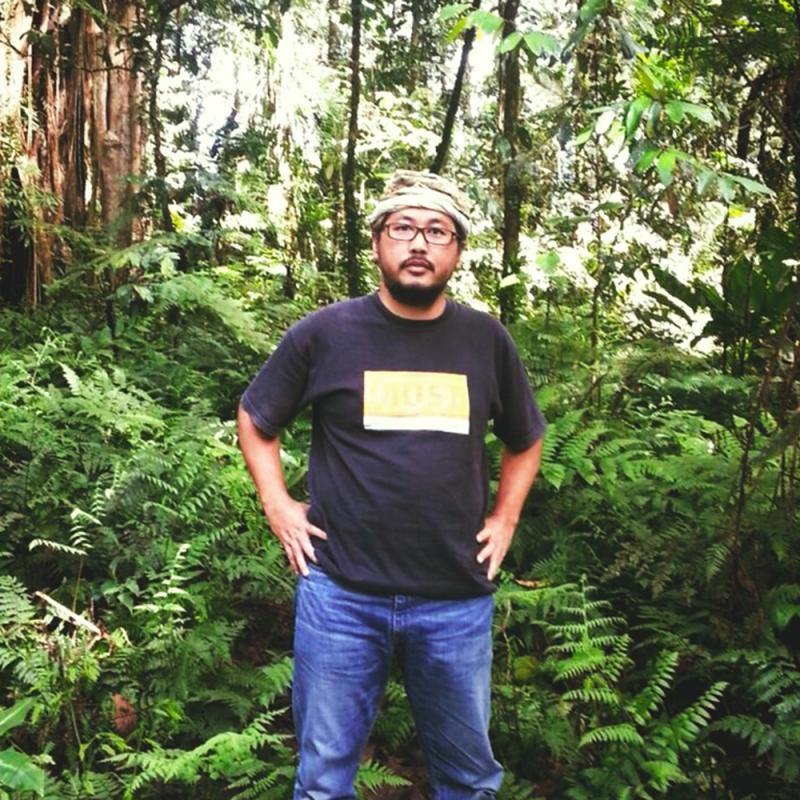Ein Mann mit Brille, schwarzem T-Shirt und Jeans steht inmitten eines dichten, tropischen Regenwaldes mit üppigem Farnbewuchs und hohen Bäumen.