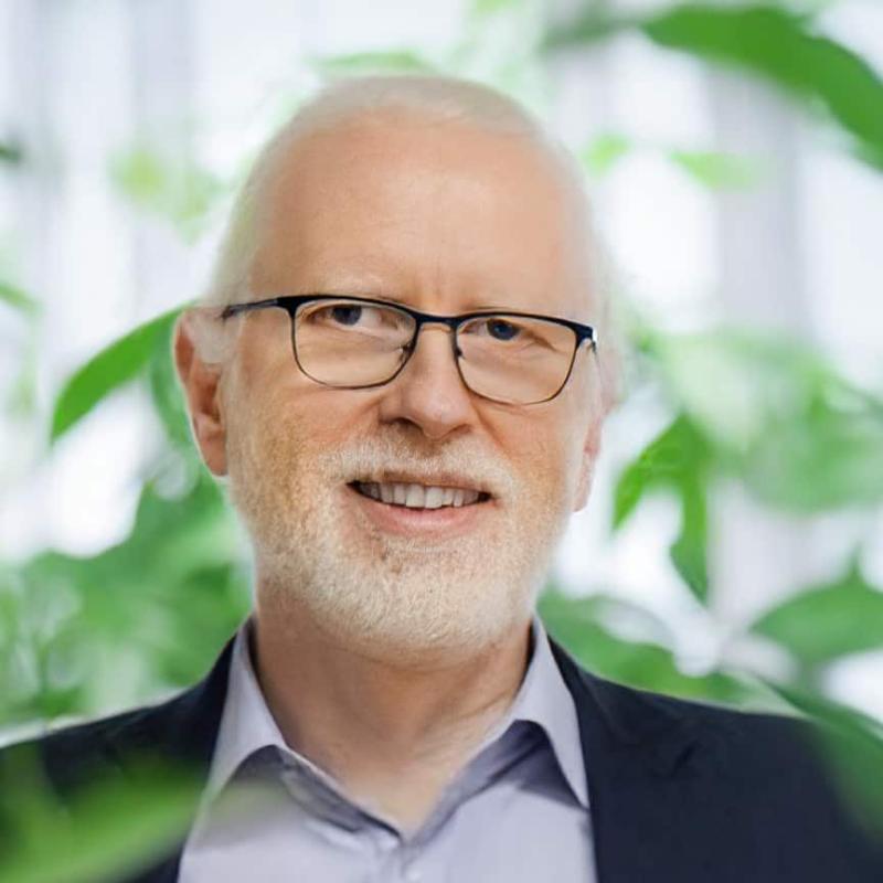 Porträt eines älteren Mannes mit weißem Bart, Brille und dunklem Sakko, umgeben von grünen Pflanzen.