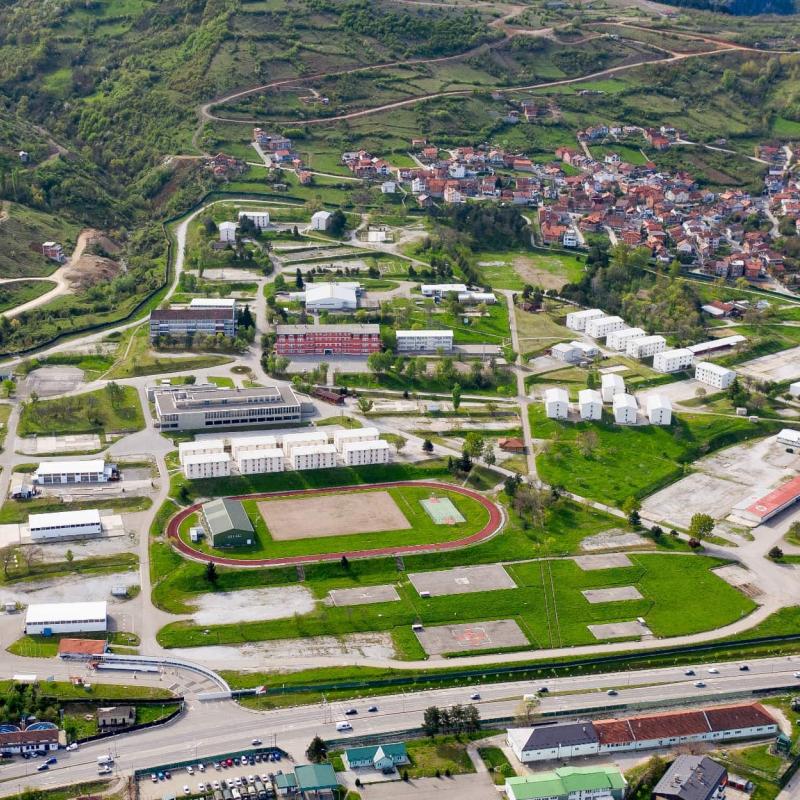 Luftaufnahme eines weitläufigen Geländes mit Gebäuden, Sportanlagen und einer angrenzenden Stadt in einer hügeligen Landschaft.