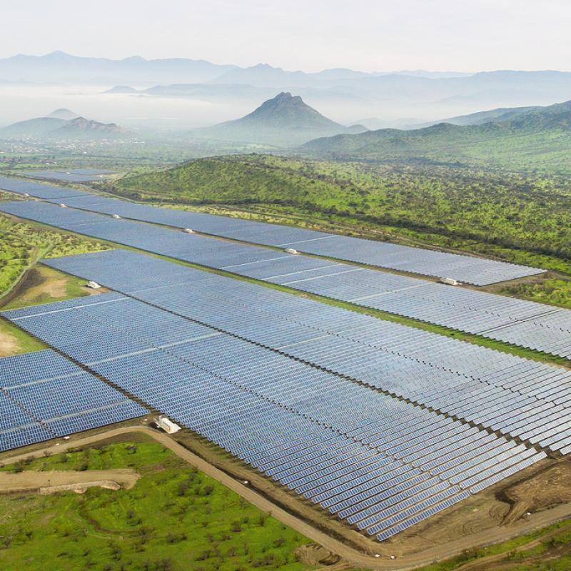 Luftaufnahme eines großen Solarparks mit zahlreichen Photovoltaik-Paneelen, umgeben von grüner Landschaft und Bergen im Hintergrund, die mit leichtem Nebel bedeckt sind.