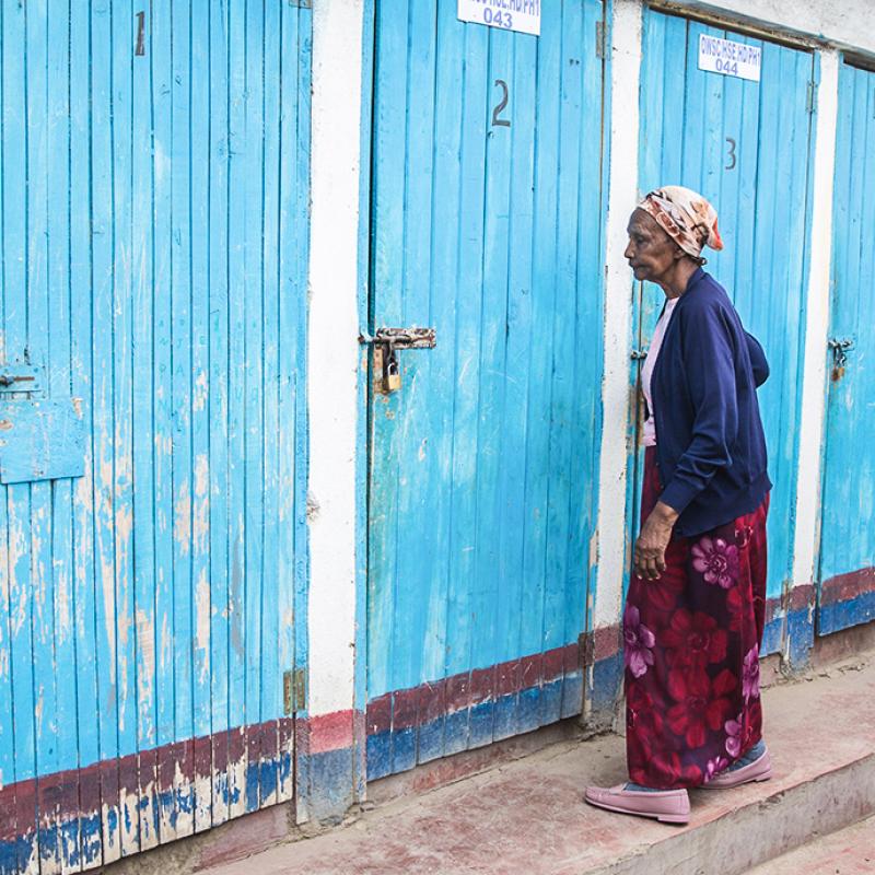 Das Bild zeigt eine Frau vor einer Reihe blauer Türen mit Nummerierungen stehend. Die Türen wirken abgenutzt mit sichtbaren Abblätterungen der Farbe und Rost an den Metallteilen. Die Frau trägt ein Kopftuch und traditionelle Kleidung.