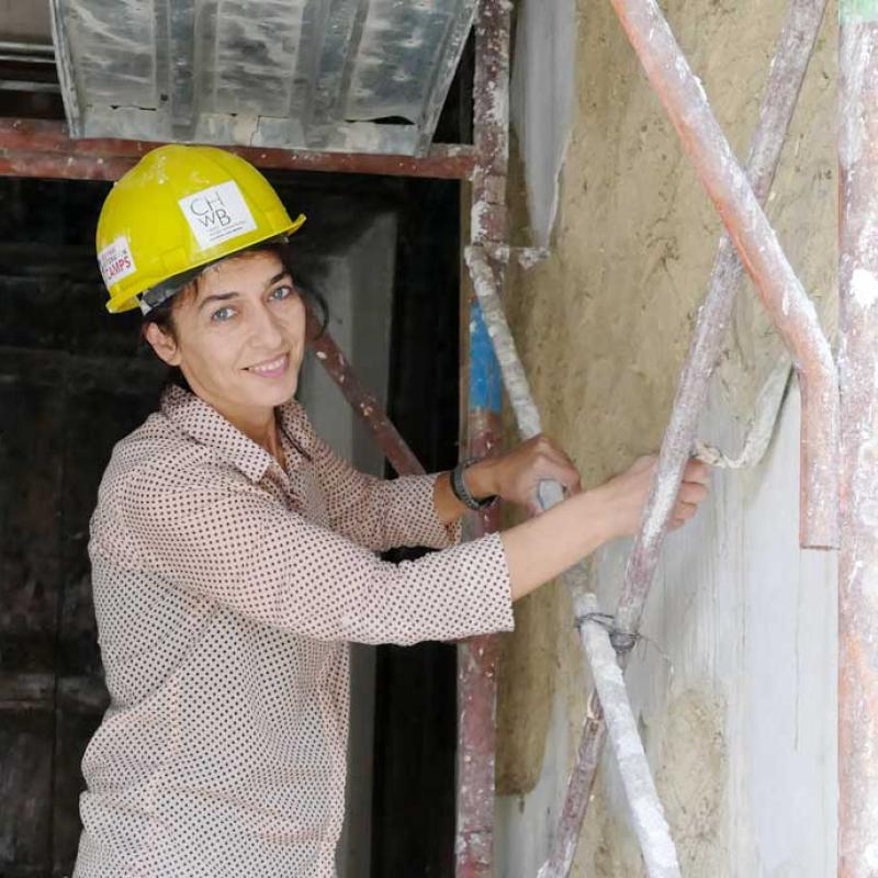 Eine circa 30 jährige Frau mit kurzen, dunklen Haaren und einem gelben Schutzhelm verputzt eine Wand in einem Raum mit roher Bausubstanz und einer alten Tür. Sie trägt ein gepunktetes Hemd und blickt lächelnd zur Kamera, während sie auf einem Gerüst steht.