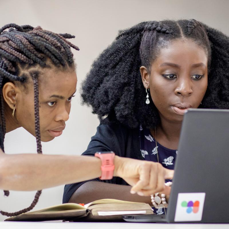 Zwei afrikanische Frauen mit dunkler Kleidung und unterschiedlichen Frisuren – die eine mit langen Zöpfen, die andere mit voluminösem, natürlichem Haar – betrachten aufmerksam einen Laptopbildschirm. Sie sitzen an einem weißen Tisch, auf dem ein Notizbuch neben dem Laptop liegt.
