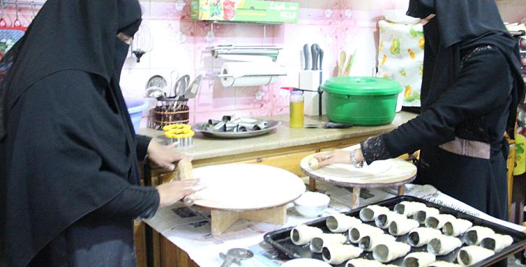Das Foto zeigt die kleine Bäckerei der 38-Jährigen Safa'a Albatool im Jemen. Sie trägt traditionelle Kleidung mit einem Hijab und bereitet gerade Essen zu.  Sie steht an einer Arbeitsplatte, auf der mehrere Backformen zu erkennen sind, und scheint Teig auszurollen. Die Küche ist mit verschiedenen Küchenutensilien und -geräten ausgestattet.