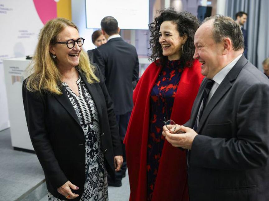 Drei Personen in Businesskleidung lachen und unterhalten sich bei einer Veranstaltung, eine Frau trägt ein schwarz-weißes Kleid, eine andere einen roten Mantel, und ein Mann trägt einen Anzug.