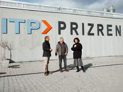 Drei Personen stehen vor einer grauen Wand mit der Aufschrift "ITP PRIZREN" und unterhalten sich.