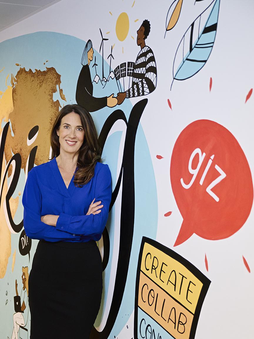 GIZ/Gaby Gerster Eine Frau mit langen braunen Haaren in einer blauen Bluse steht mit verschränkten Armen vor einer bunten Wand mit der Aufschrift „giz“ und Illustrationen zu Nachhaltigkeit und Zusammenarbeit.